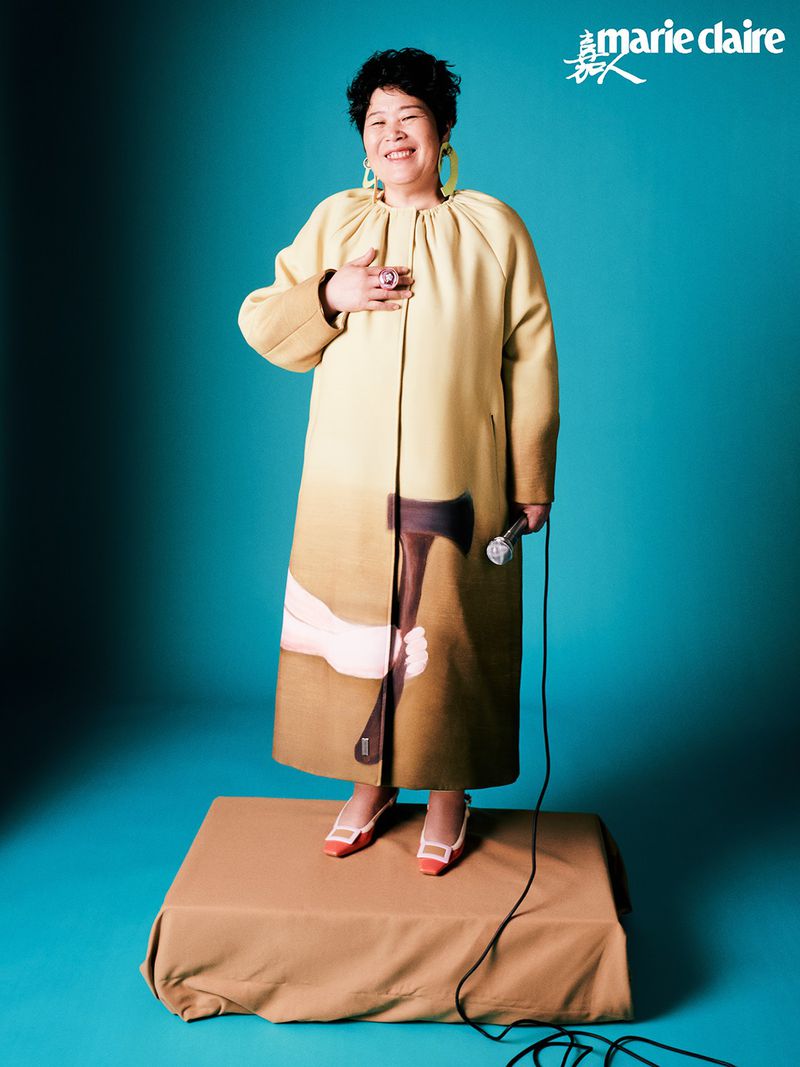 Editorial shot of a smiling Asian woman in a patterned coat, posing on a platform with a microphone in hand, set against a blue background.