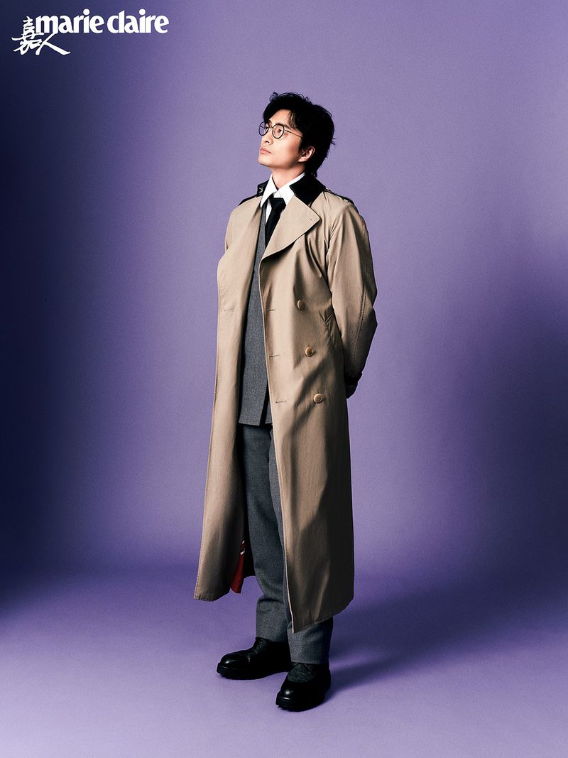 East Asian man wearing glasses and a long beige trench coat over a grey suit standing against a purple studio backdrop.