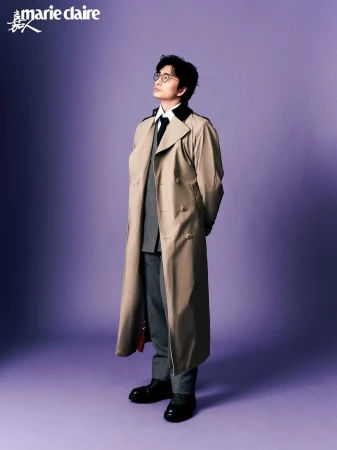 East Asian man wearing glasses and a long beige trench coat over a grey suit standing against a purple studio backdrop.