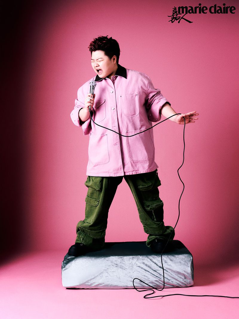 Adult male wearing a pink shirt and green pants stands on a platform holding a microphone against a pink studio background. 
