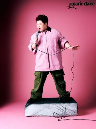 Adult male wearing a pink shirt and green pants stands on a platform holding a microphone against a pink studio background. 