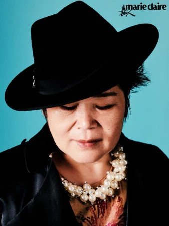 Close-up studio shot of a stylish adult woman in a black hat and pearl necklace against a blue background, looking downwards with neutral expression.