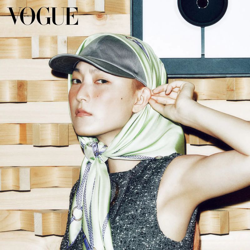 Headshot of a woman in a baseball cap and scarf with a wood-block background. Vogue logo visible. Fashion editorial style.