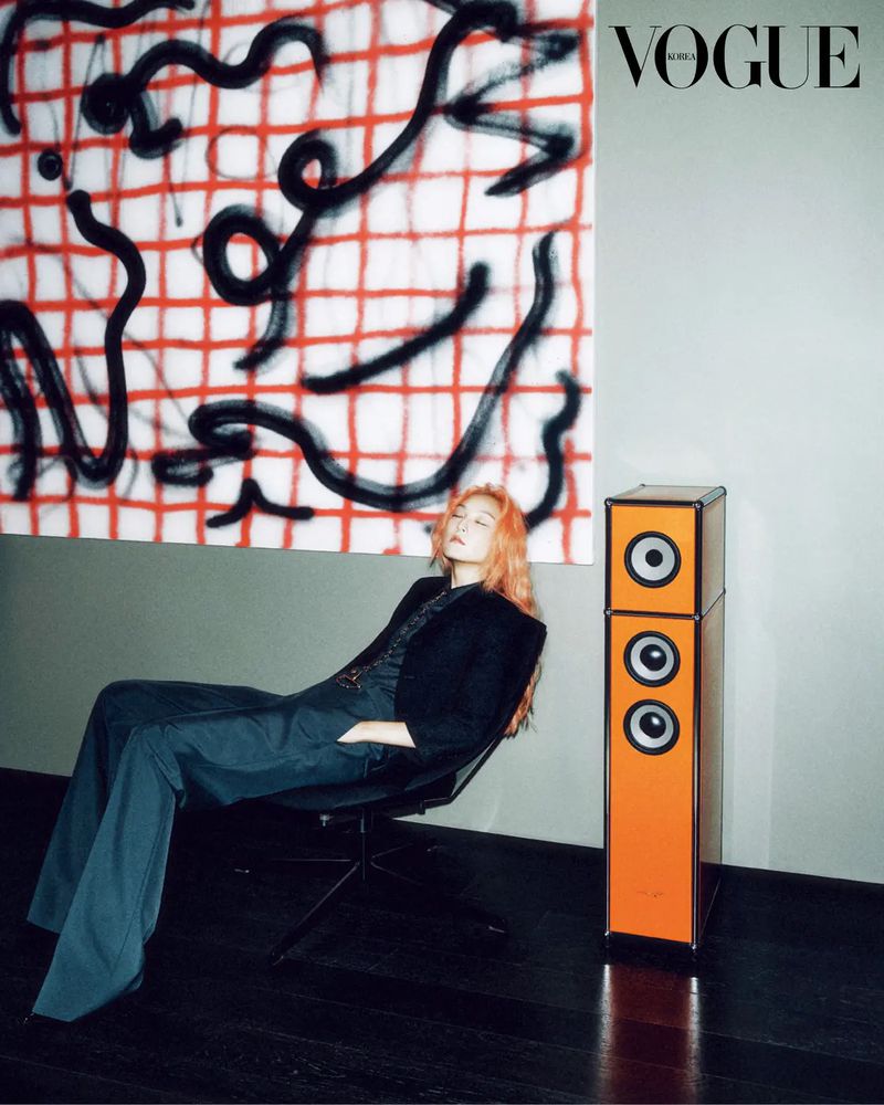 A fashion shot features a woman with orange hair in a black blazer and wide leg pants, seated next to an orange speaker and graphic art.