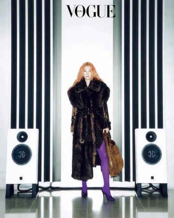 Editorial fashion shot: woman in long fur coat, purple tights, and speakers, with 'Vogue Korea' logo and black-and-white striped background.