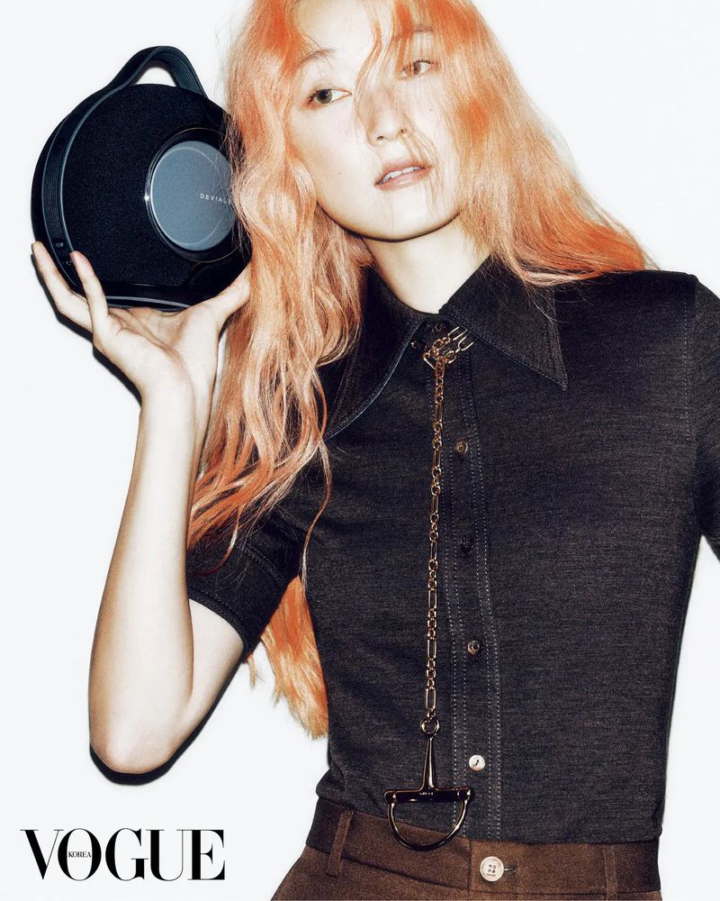 Woman with peach hair poses in studio with a black speaker. She wears a collared shirt with a chain and dark brown pants.
