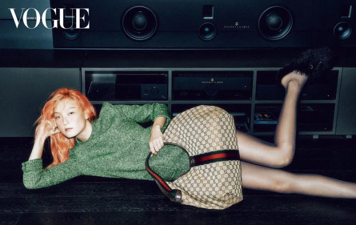 Fashion editorial featuring a woman with orange hair in a green dress reclining on a dark floor with a Gucci handbag.