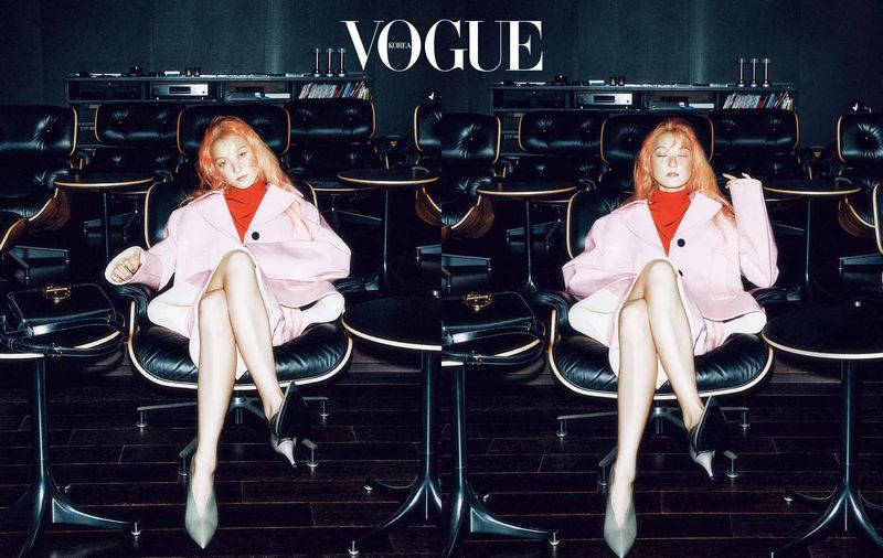 Fashion shot featuring an adult woman in a pink coat sitting in a black chair, complemented by a black purse, set against a dark indoor backdrop.