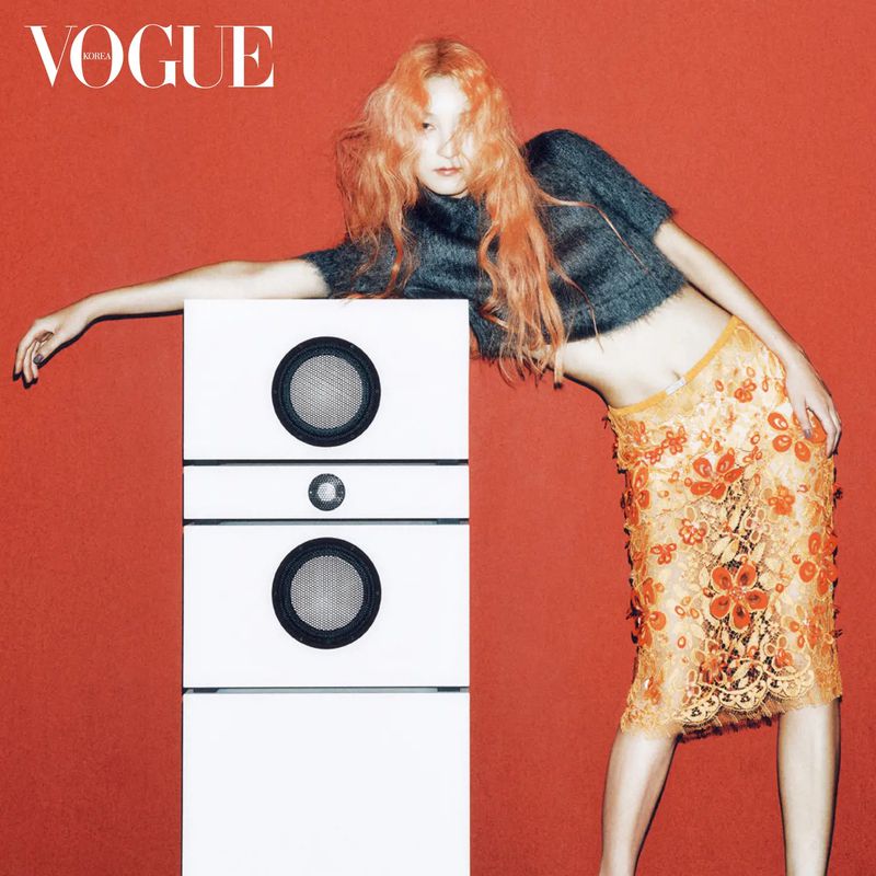 Woman with orange hair poses in a gray sweater and floral skirt next to a white speaker on a red background for a magazine cover.