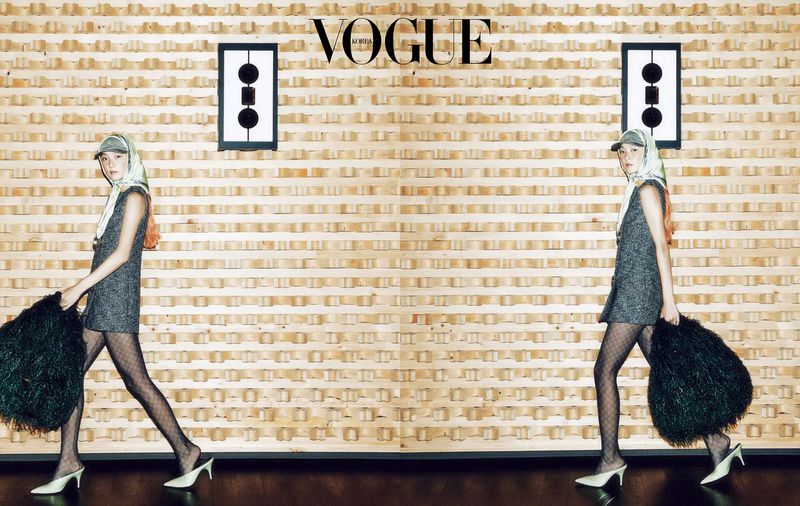 A young woman poses in a studio wearing a dress, heels and headscarf, holding a large black feathered bag against a decorative wall.
