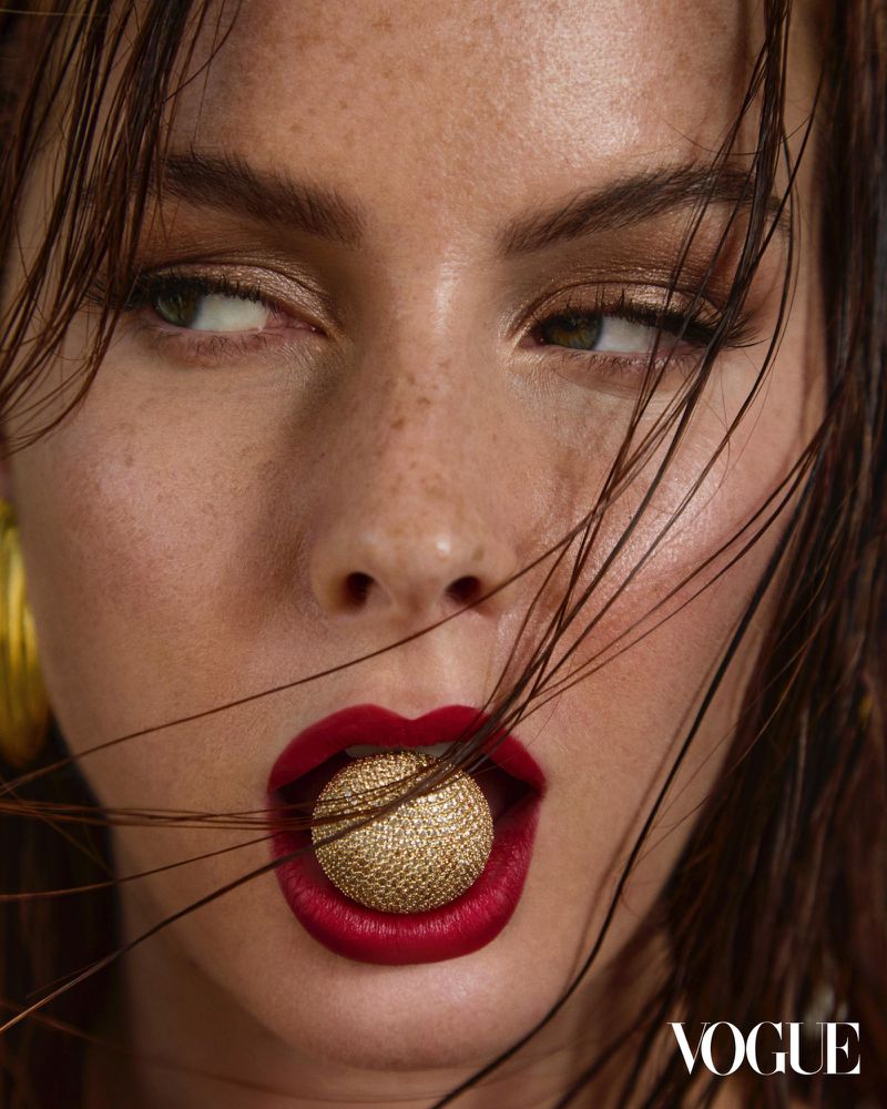 Close-up portrait of a woman with red lipstick holding a gold sphere in her mouth, wet hair, and gold earrings, Vogue logo.