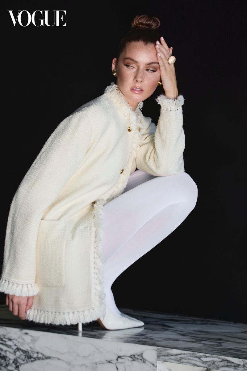 A woman in a cream-colored jacket poses in a studio with a black background, kneeling on a marble surface, wearing white tights and heels.