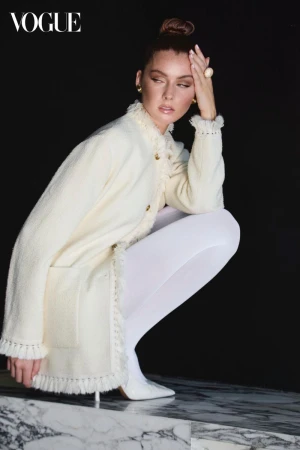 A woman in a cream-colored jacket poses in a studio with a black background, kneeling on a marble surface, wearing white tights and heels.