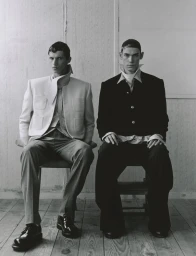 Two male models in sculptural high-collar tailoring seated side by side on wooden chairs against a panelled wall.