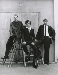 Three young male models posing on wooden ladders wearing dark suits and platform boots against a rustic concrete wall.