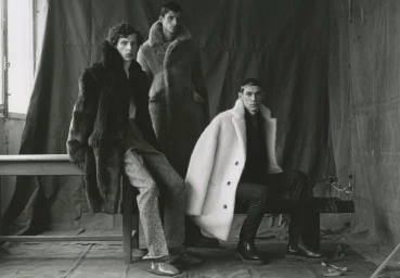 Three male models wearing textured winter overcoats and tailored trousers posed in a monochrome studio setting with fabric drapery.