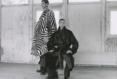 Two male models posing indoors, one seated in a dark belted coat and one standing behind him in an oversized zebra print coat.