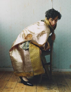 Model crouching on a rustic wooden chair wearing an oversized metallic gold coat with textured trim and black pointed shoes.