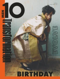 Model wearing a metallic gold coat and black shoes, leaning on a wooden chair indoors.
