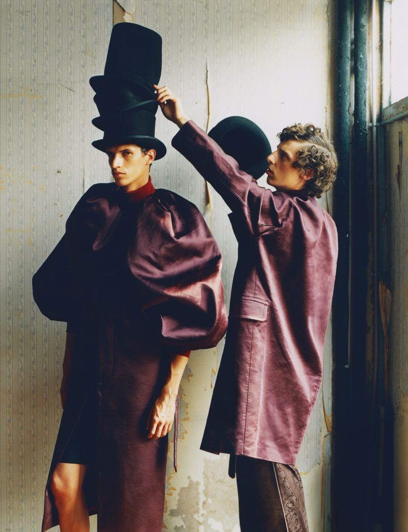 Two young men in a fashion-forward image. One man wears stacked hats while the other arranges a smaller hat. The background is a distressed wall.