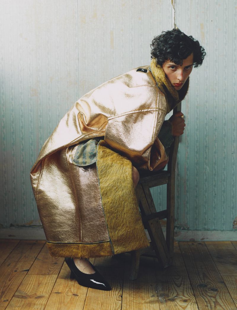 Adult female wearing a metallic gold and fur coat, seated on a wooden ladder inside a room with patterned walls and wood floors.