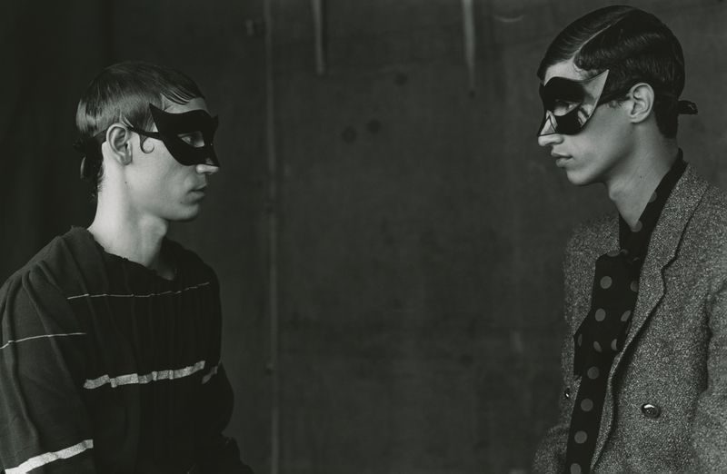 Two young adult males in an indoor setting, wearing black masquerade masks and stylish attire, facing each other against a dark backdrop.