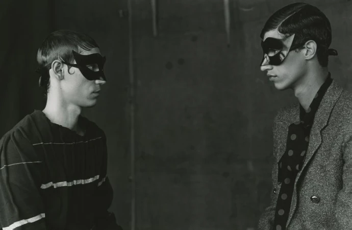 Two young adult males in an indoor setting, wearing black masquerade masks and stylish attire, facing each other against a dark backdrop.
