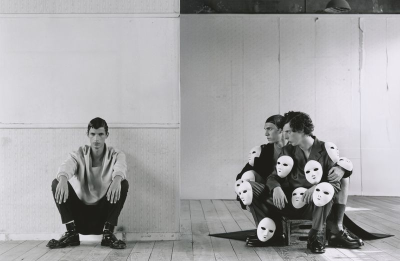 Monochrome image featuring one man squatting against a white wall and three men huddled on a crate surrounded by face masks.