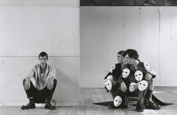 Monochrome image featuring one man squatting against a white wall and three men huddled on a crate surrounded by face masks.