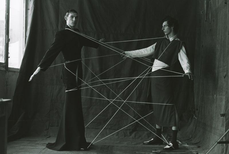 Monochromatic studio portrait of two slender adult men connected by string, wearing dark clothing, in a minimalist setting. 