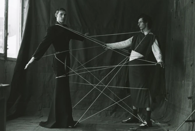 Monochromatic studio portrait of two slender adult men connected by string, wearing dark clothing, in a minimalist setting. 