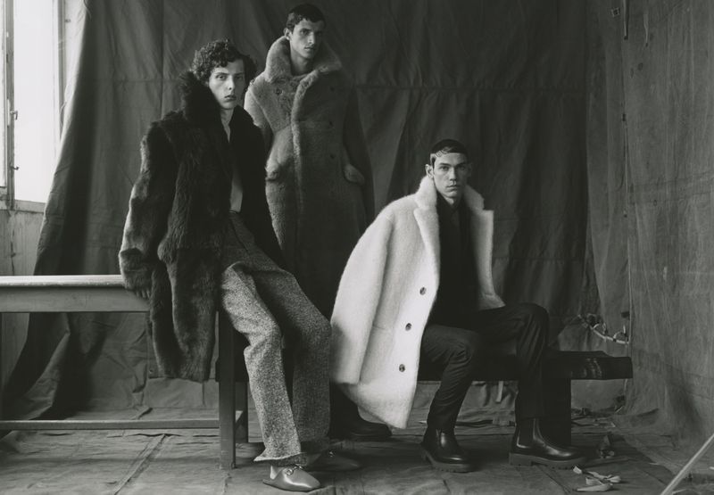 Three male fashion models pose indoors wearing faux fur coats in an eye-level shot. One model sits, while two are standing in the studio.