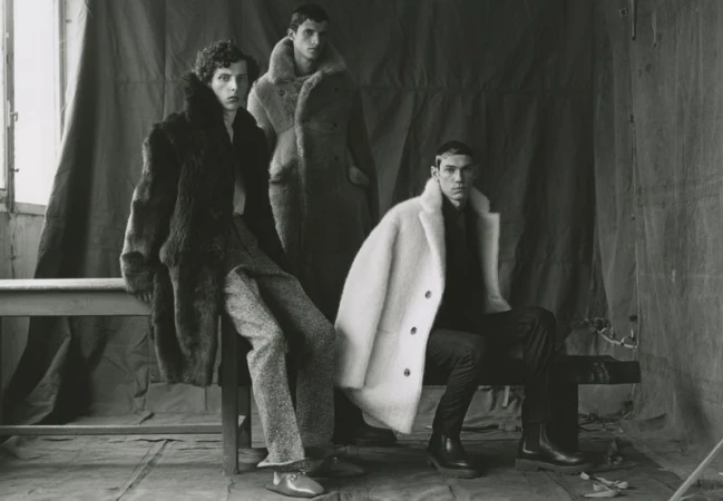 Three male fashion models pose indoors wearing faux fur coats in an eye-level shot. One model sits, while two are standing in the studio.