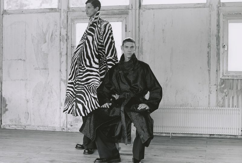 Two adult men in a sparse room with white walls, one wearing a zebra-print coat, the other a patterned coat, for a fashion editorial.