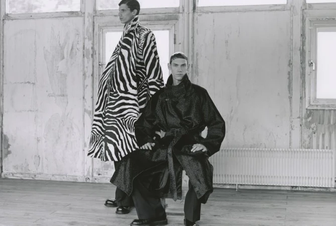 Two adult men in a sparse room with white walls, one wearing a zebra-print coat, the other a patterned coat, for a fashion editorial.