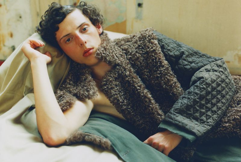 Young adult lies on a light green bed wearing a brown sheepskin coat with a quilted gray jacket sleeve in an indoor setting.
