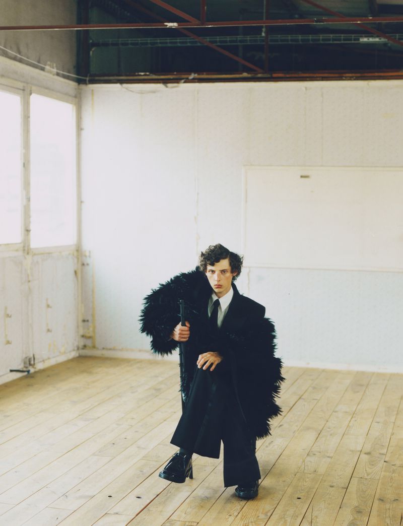A young adult poses in a black suit and furry coat in an empty room with a wood floor and bright window light.