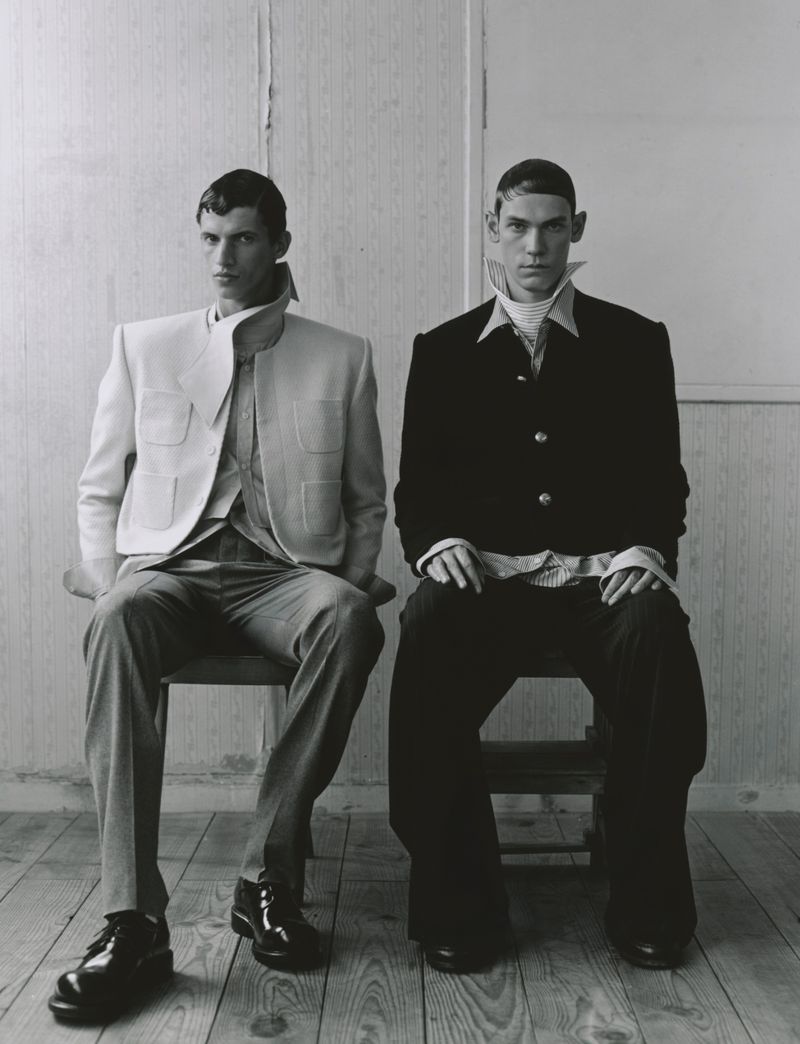 Black and white portrait of two men sitting on wooden chairs, wearing jackets, dress pants, and shoes in an indoor environment.