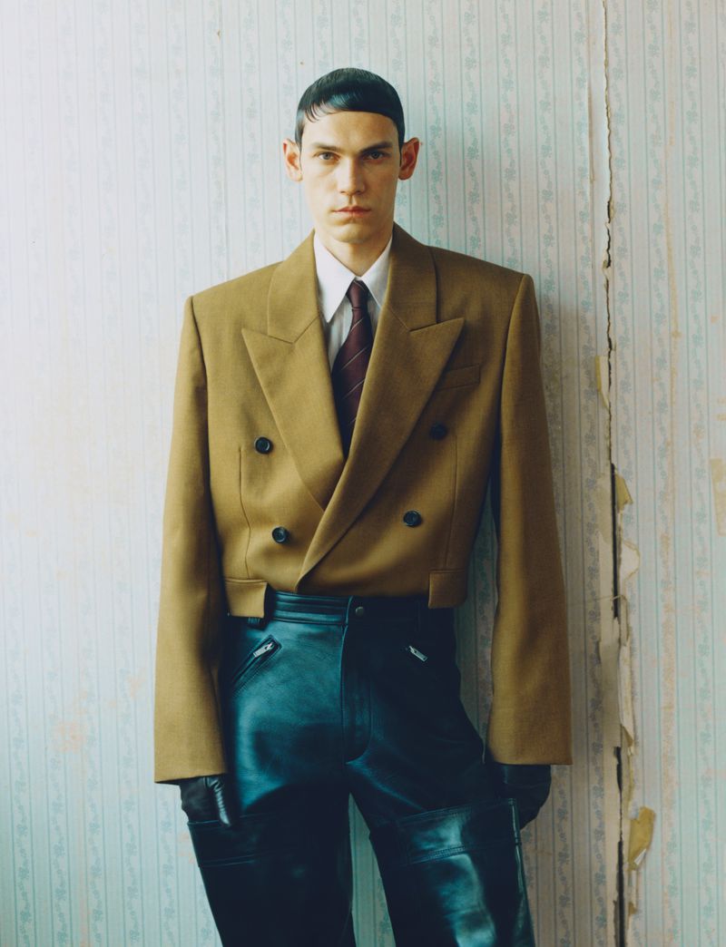 Portrait of a slender adult male in a cropped brown blazer and leather pants, posing against a vintage wallpaper background indoors.