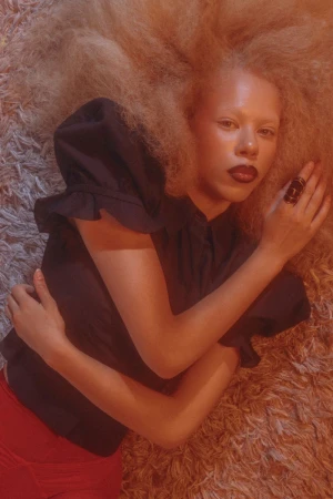 Woman with fair skin and dark lipstick lies on a textured rug with a dark blouse and large hair. Fashion editorial portrait.