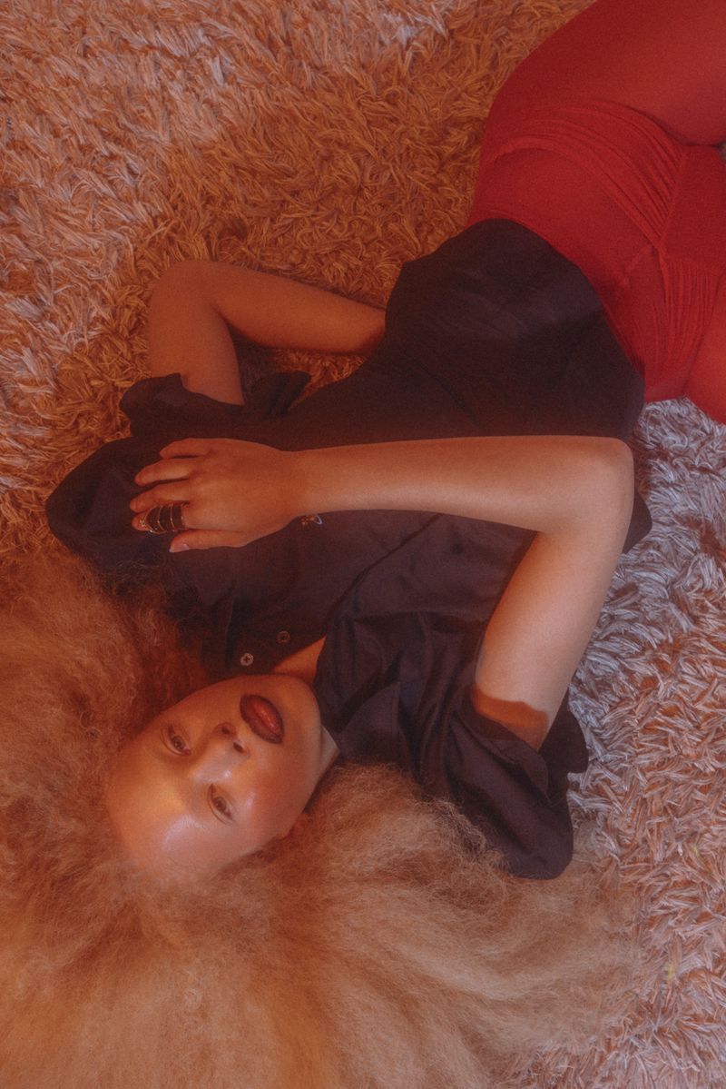 Woman with blonde hair in a dark shirt and red pants lies on a shaggy carpet looking at the camera in a portrait orientation.