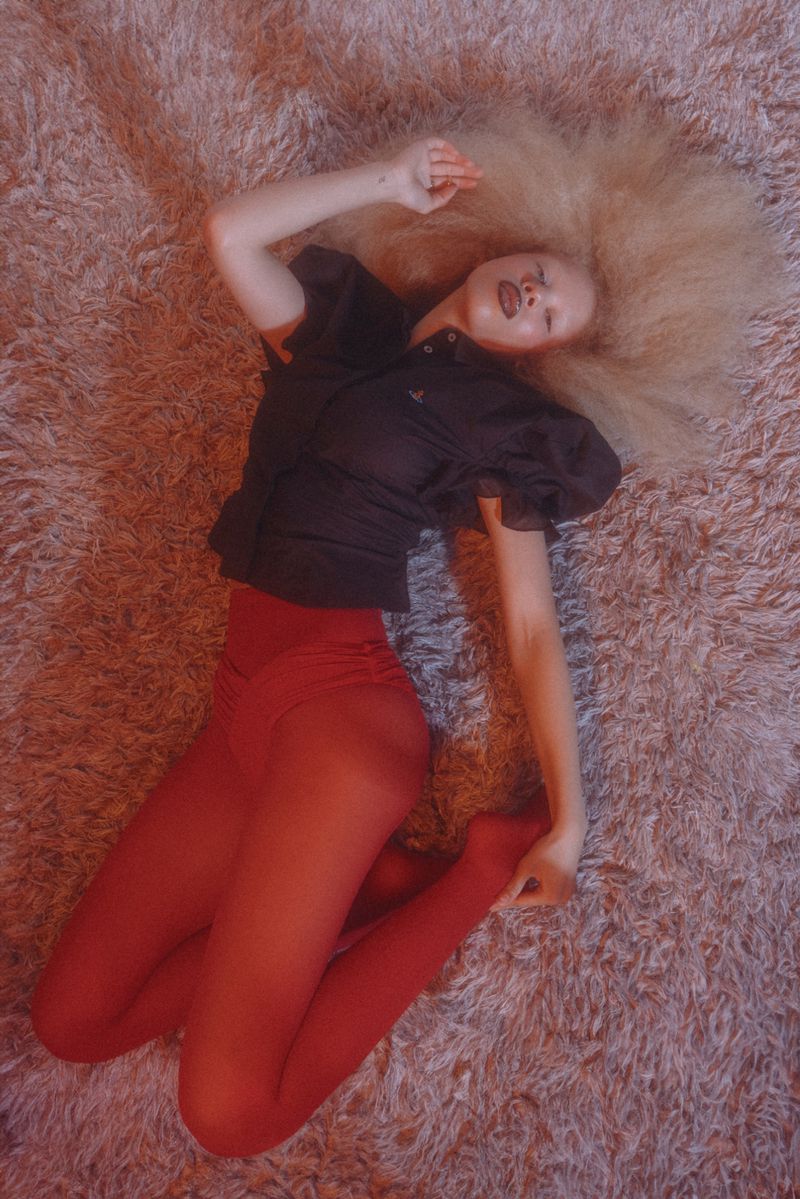 Overhead shot of a fair-skinned adult female with blonde Afro hair, wearing a dark top and red tights, lying on a fuzzy light-pink rug.