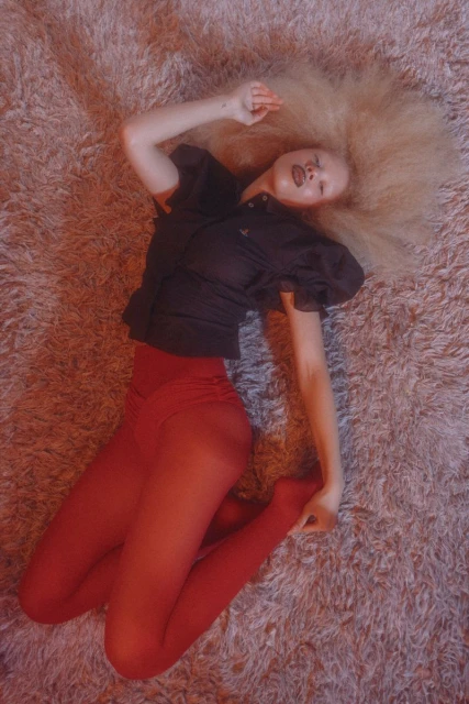 Overhead shot of a fair-skinned adult female with blonde Afro hair, wearing a dark top and red tights, lying on a fuzzy light-pink rug.