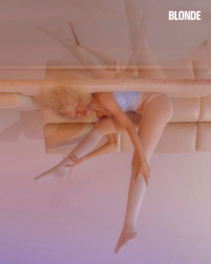 A woman with blonde hair, wearing a blue top and white hosiery, poses in a photo studio with soft lighting and multiple exposure effect.