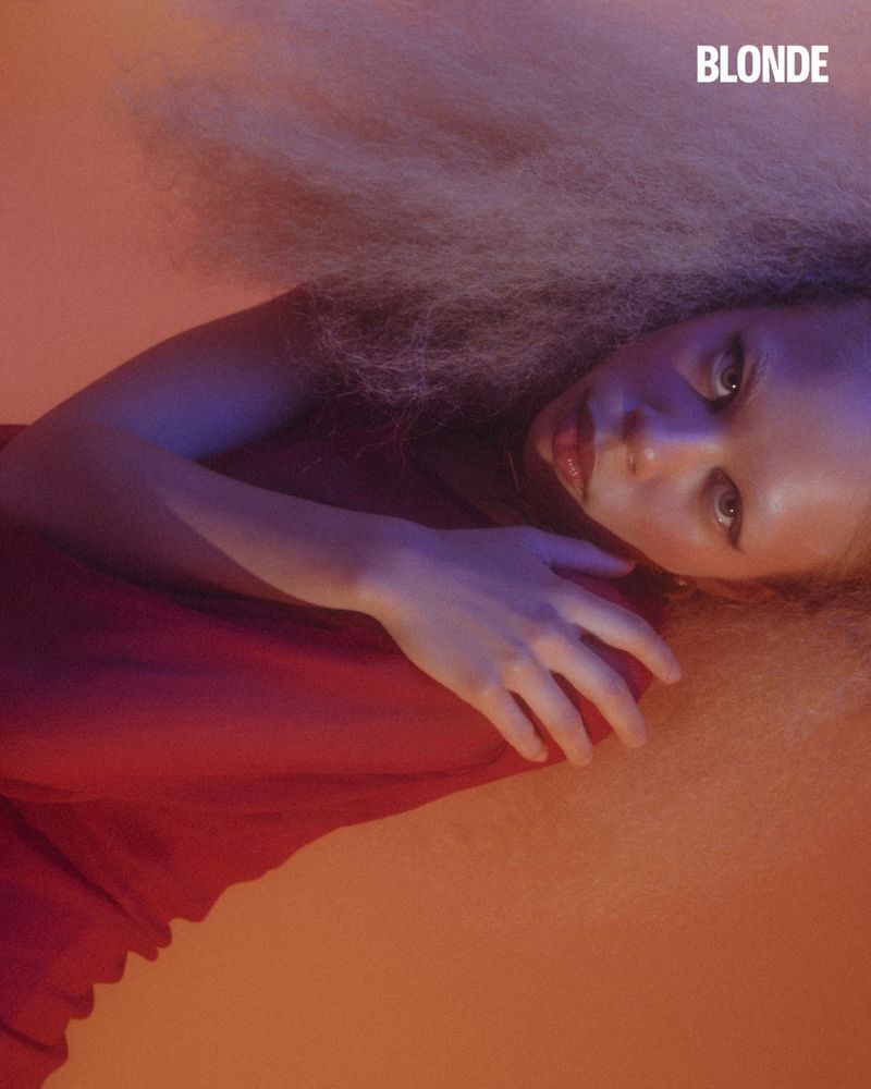 Close-up portrait of a young woman with blonde hair and red lipstick, lying down against an orange backdrop in a studio setting.