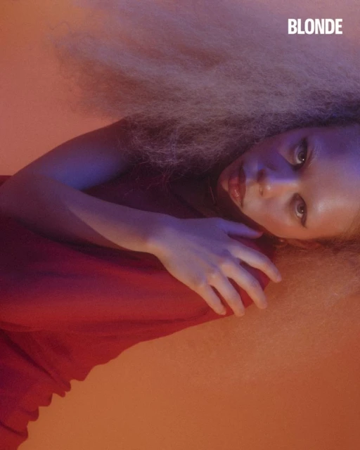 Close-up portrait of a young woman with blonde hair and red lipstick, lying down against an orange backdrop in a studio setting.