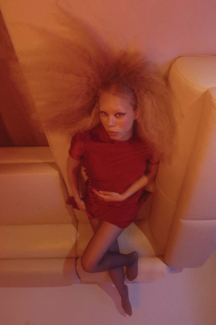 Eye-level shot of a fair-skinned female in a red dress lying on a cream-colored couch with bright lighting and an afro hairstyle.