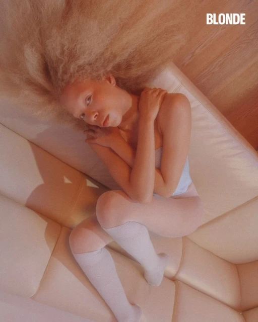 Overhead shot of a woman with blonde hair lying on a white couch, wearing white socks, and a white top in a relaxed pose.
