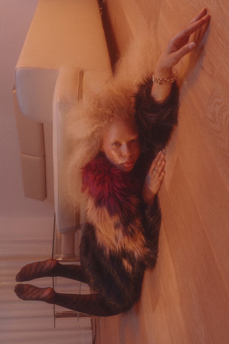 Stylish woman in a colorful fur coat reclines on a wooden floor next to a cream sofa. The lighting is warm and atmospheric.