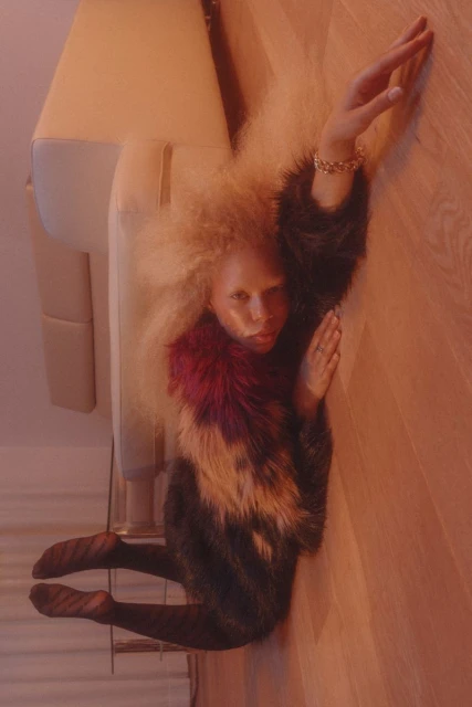 Stylish woman in a colorful fur coat reclines on a wooden floor next to a cream sofa. The lighting is warm and atmospheric.